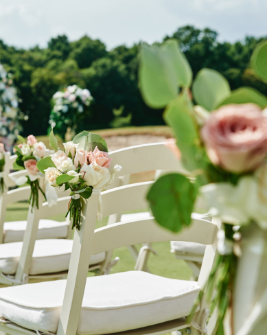 Elegant outdoor ceremony chairs decorated with fresh roses and greenery, set on a sunny lawn with trees in the background