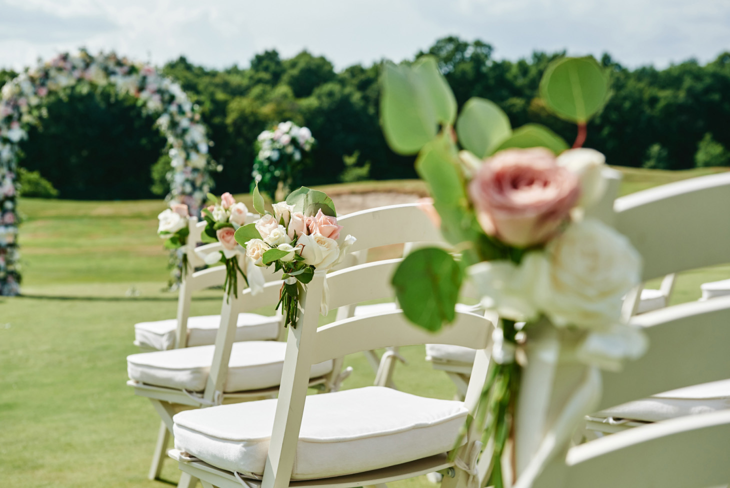 Outdoor wedding setup with white chairs, pink and white roses, semi-circular layout, greenery, and trees in the background