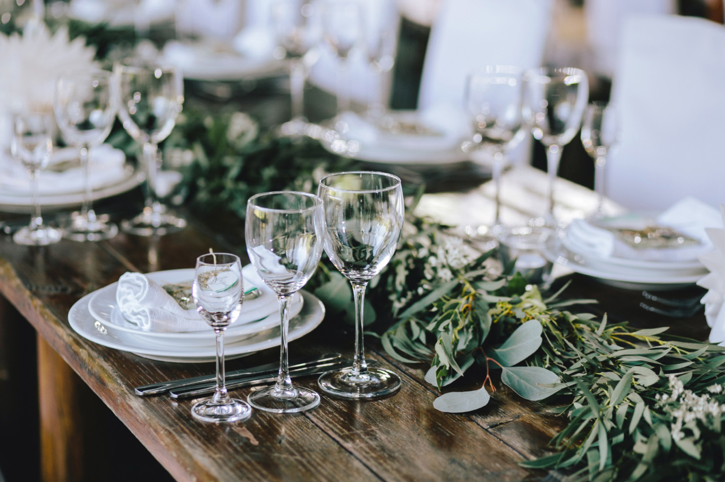 Rustic banquet table set with plates, napkins, empty wine glasses, and eucalyptus greenery as centerpiece