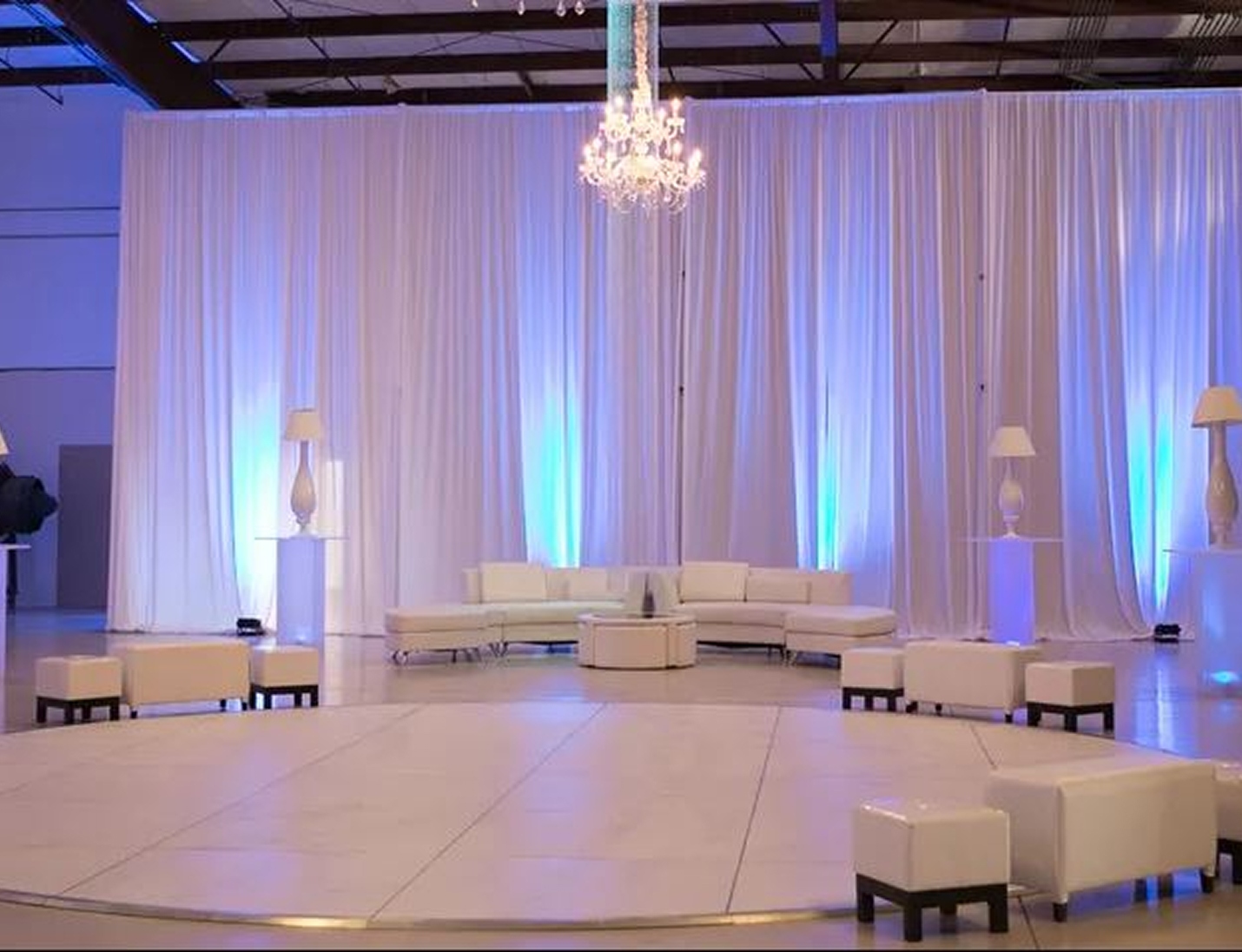 Sun ‘n Fun - Modern event space with white lounge furniture, tall draped curtains, blue uplighting, and chandeliers