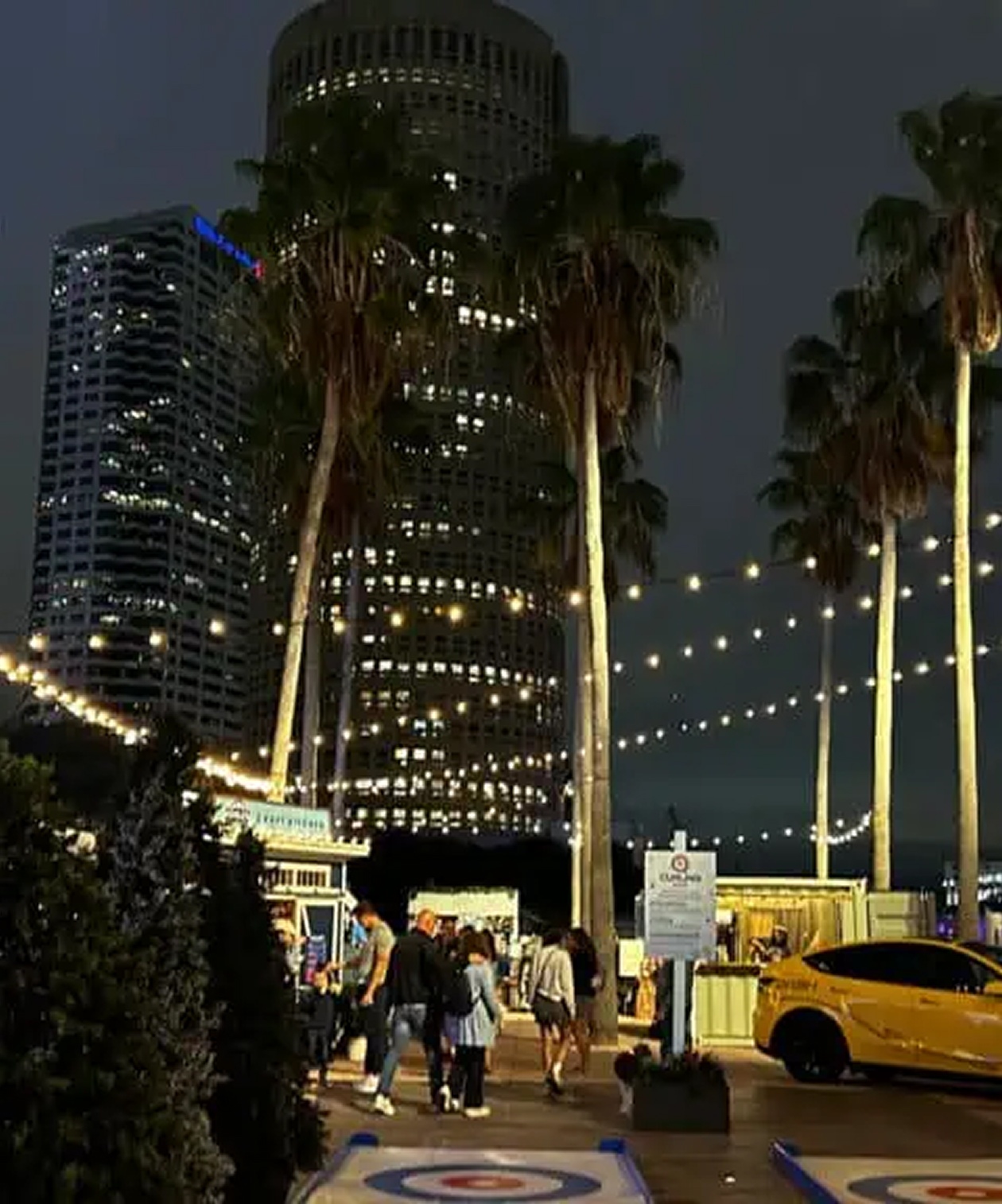 Nighttime scene at Curtis Hixon Waterfront Park with palm trees, market lights, and people walking by downtown buildings