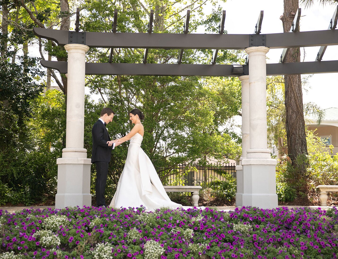 Bride and groom holding hands under a stone pergola surrounded by greenery and purple flowers at Bonnet Springs Park