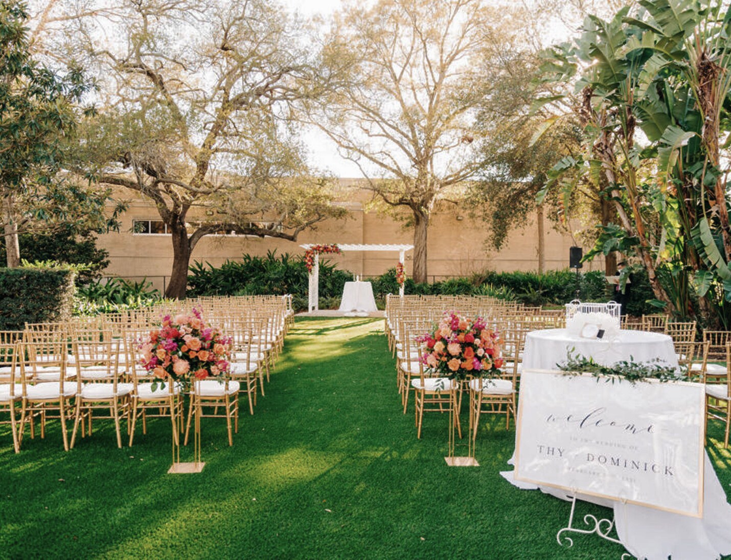 Outdoor wedding ceremony at Tampa Garden Club with gold chairs, floral aisle arrangements, and a white arch under trees