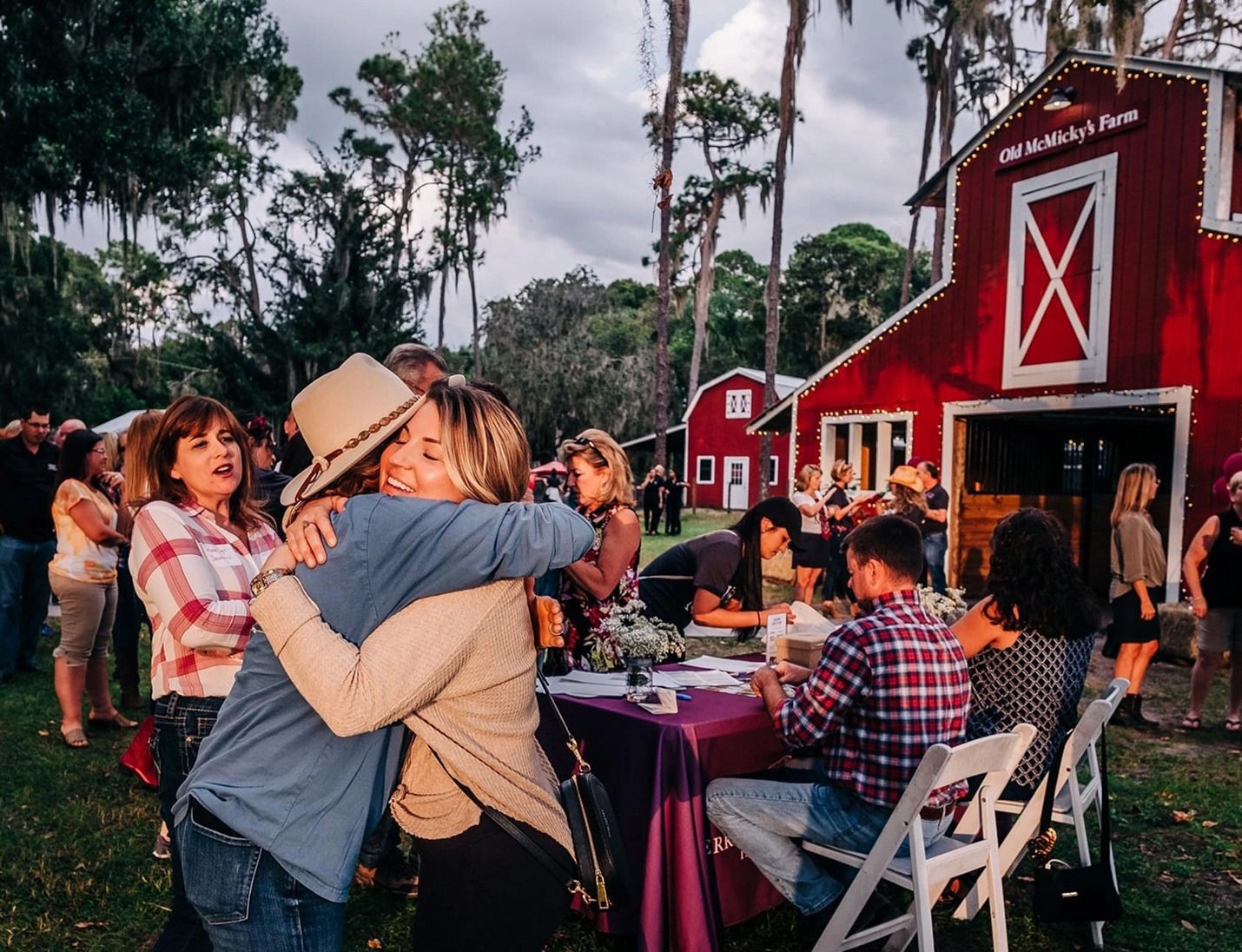 Outdoor gathering at Old McMicky’s Farm with guests hugging and mingling near the red barn under evening lights