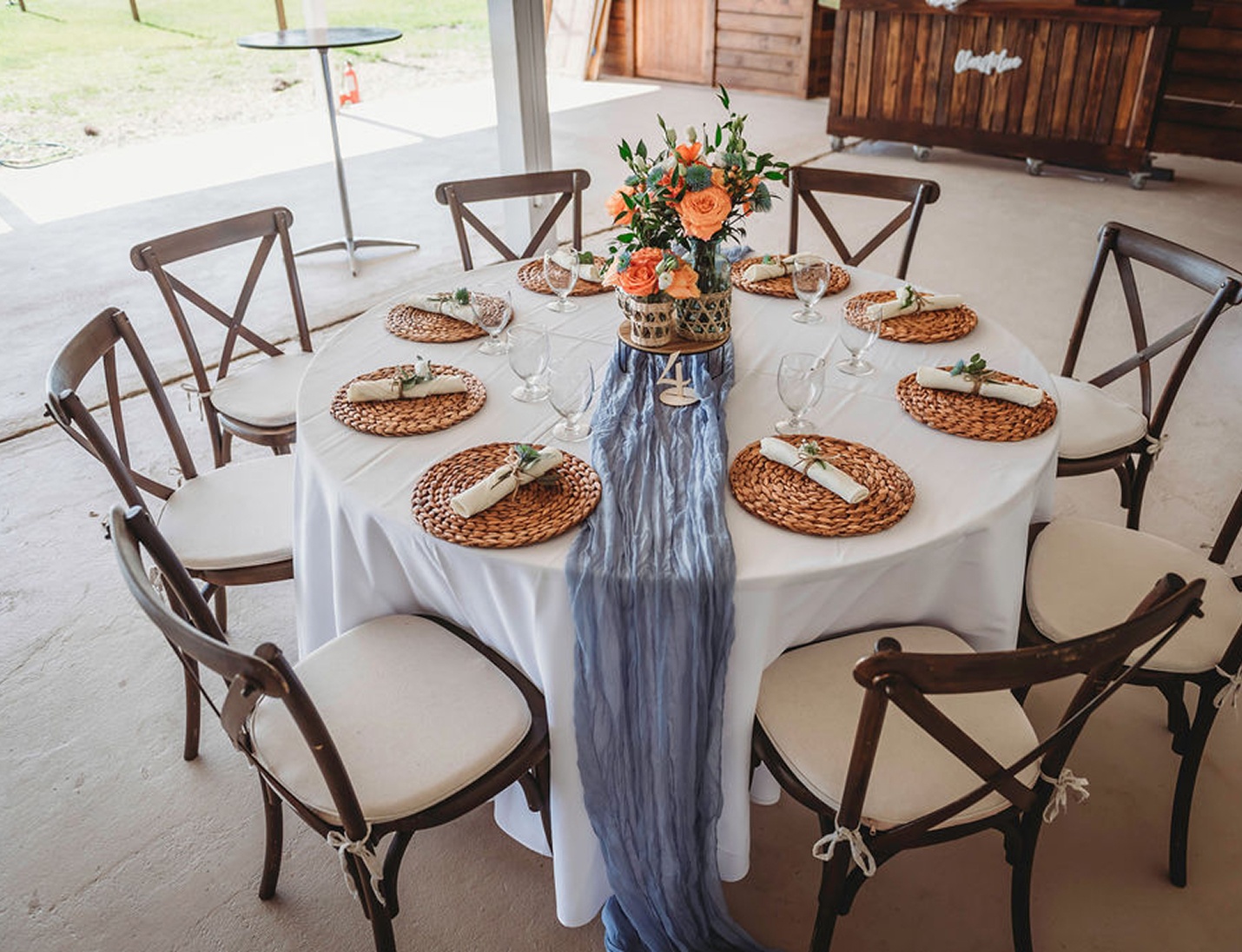Rustic table setup at Happy Hearts Barn featuring woven chargers, orange floral centerpiece, and blue table runner