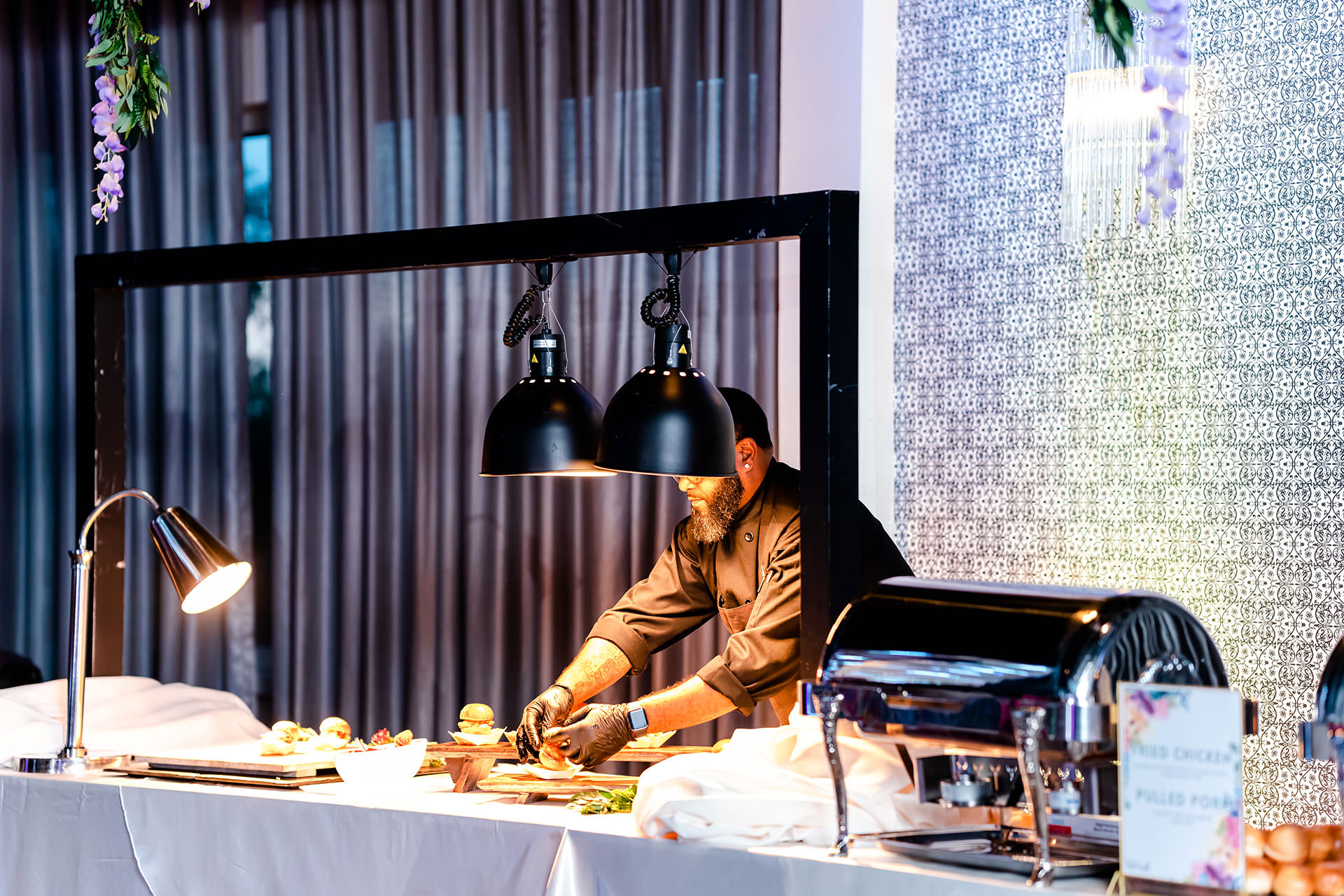 Chef preparing food under black heat lamps at The MEZZ, part of a live catering station with a modern industrial setup