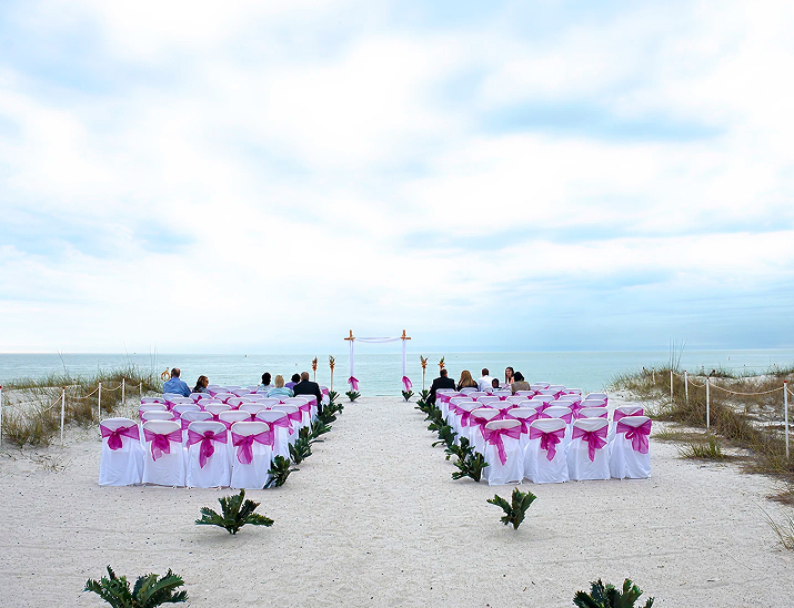 Beach wedding ceremony at Lions Club Beach House – St. Petersburg with white chairs, pink bows, and ocean backdrop