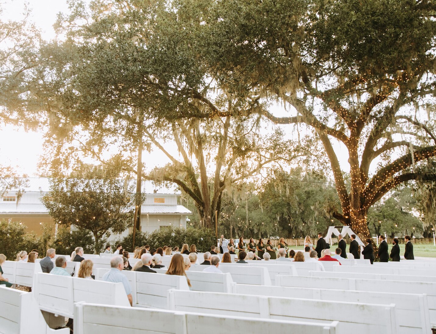 Outdoor ceremony at Ever After Farms with white benches, tree lights, and guests seated under the warm sunset glow