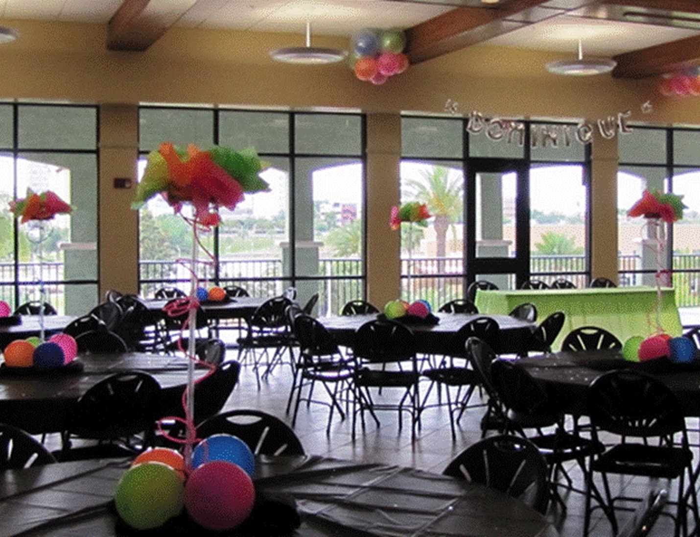 Colorful indoor setup at The Peggy Brown Building with black tables, balloons, and large windows overlooking palm trees