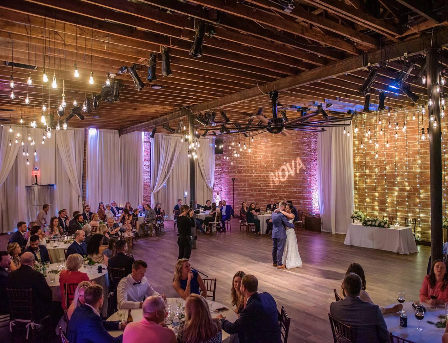 Indoor wedding reception at Nova 535 with string lights, brick walls, and a couple dancing surrounded by guests