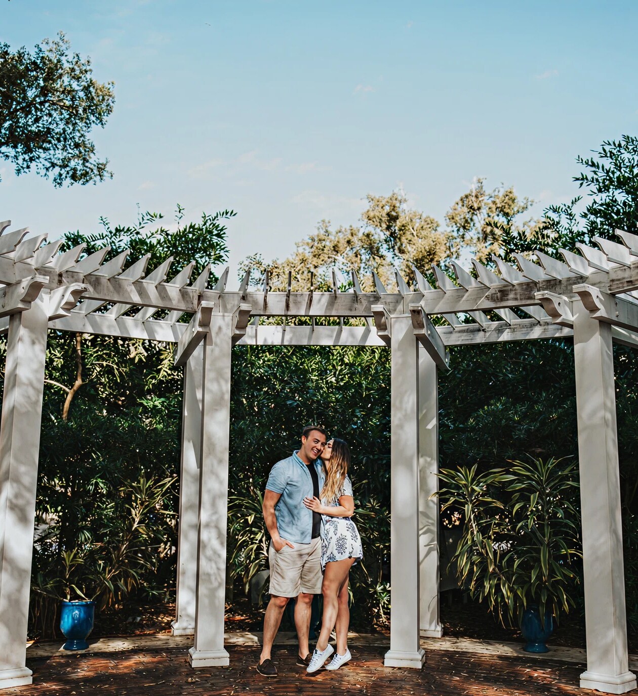 A couple sharing a tender moment beneath a white pergola surrounded by lush greenery at Harry P. Leu Gardens