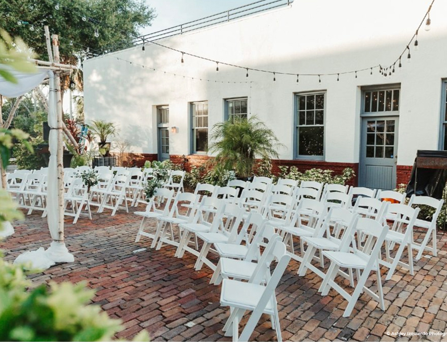 Red Door No. 5 courtyard wedding setup with white folding chairs, brick flooring, string lights, and greenery accents
