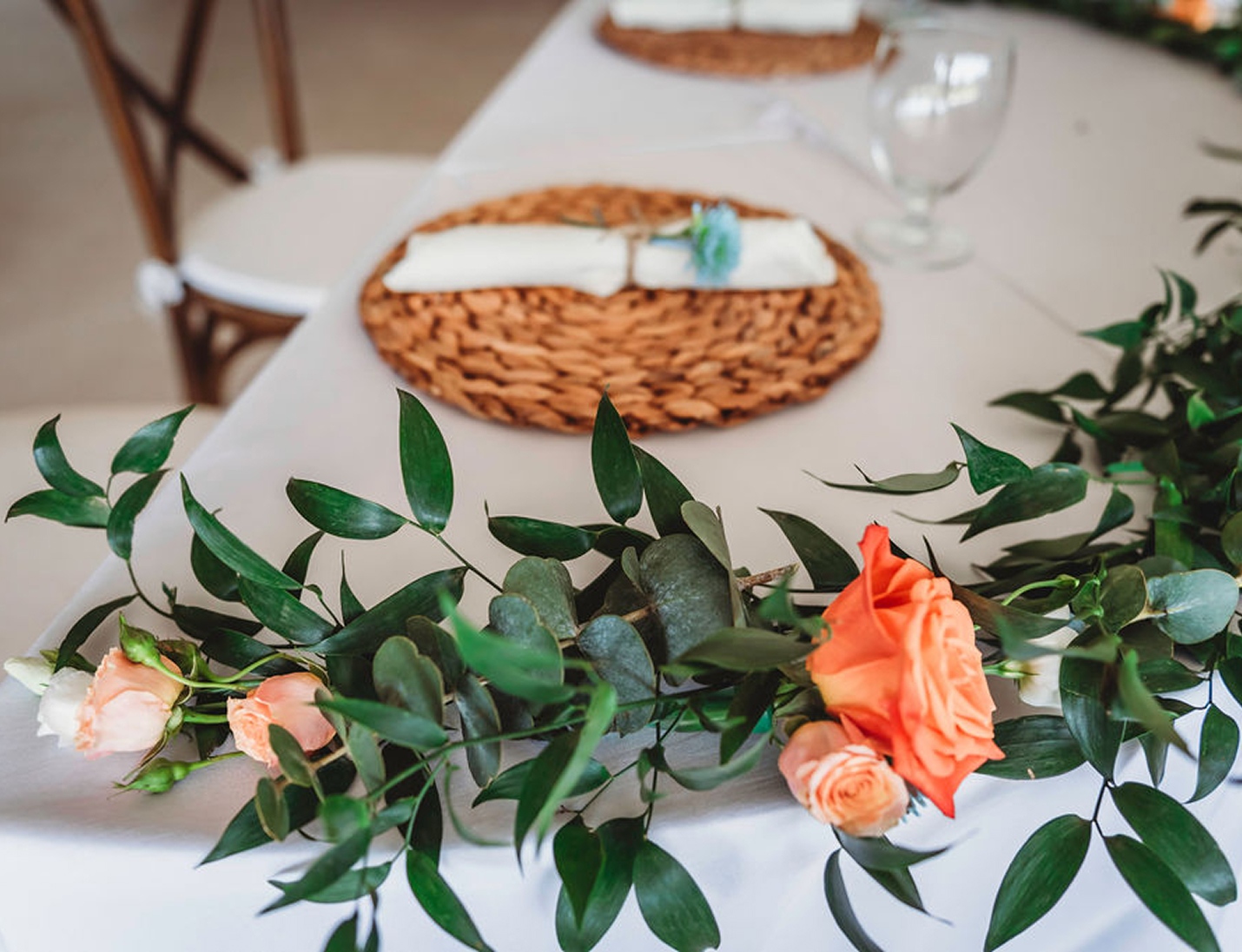 Table decor with a woven charger, a white napkin, and an orange rose greenery garland at Happy Hearts Barn reception