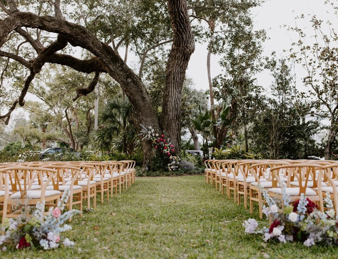 Ceremony setup at Victoria 1883 with wooden chairs, lush greenery, and floral arrangements beneath a large oak tree