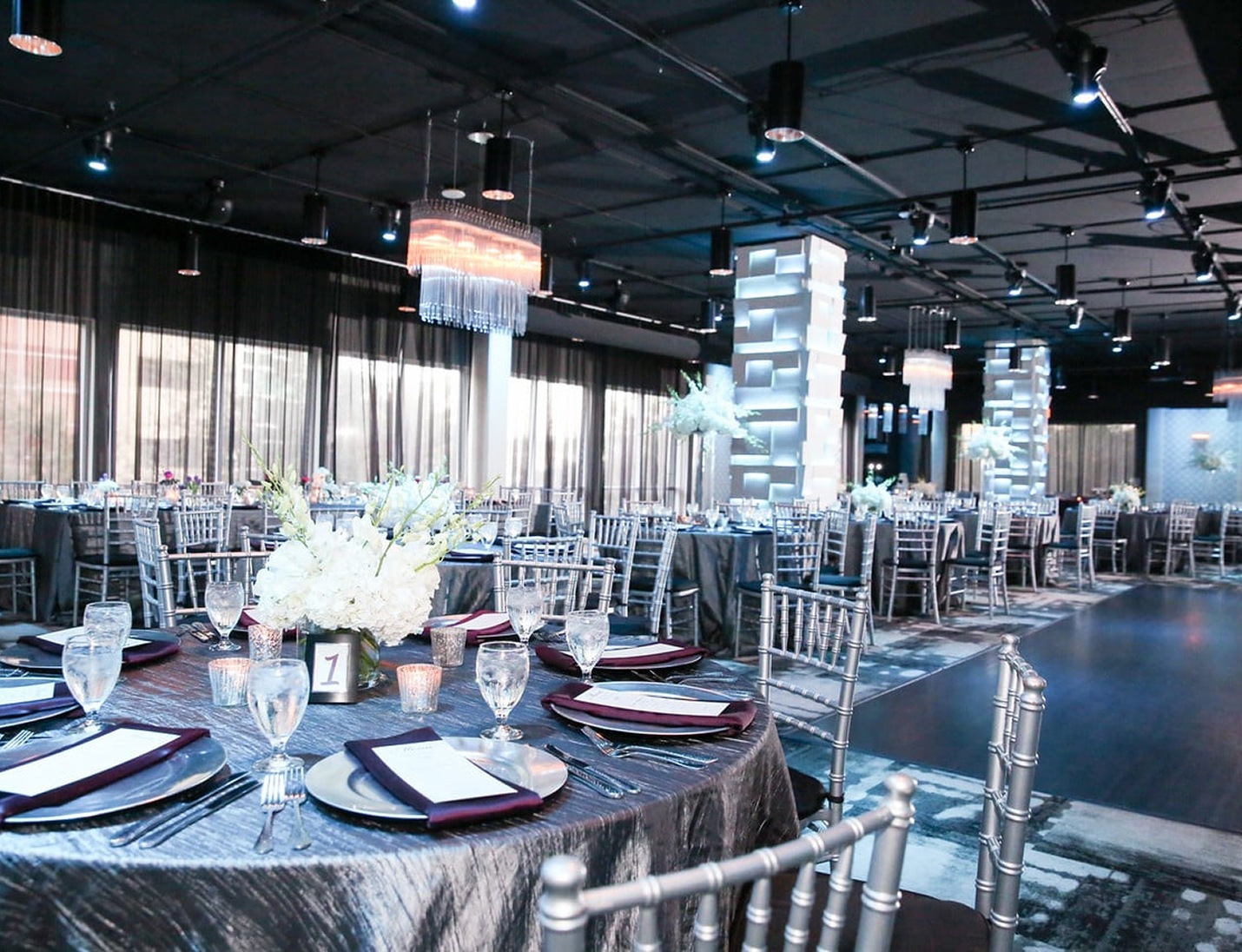 Elegant reception setup with silver chairs, draped tables, soft blue lighting, and modern chandeliers at The MEZZ