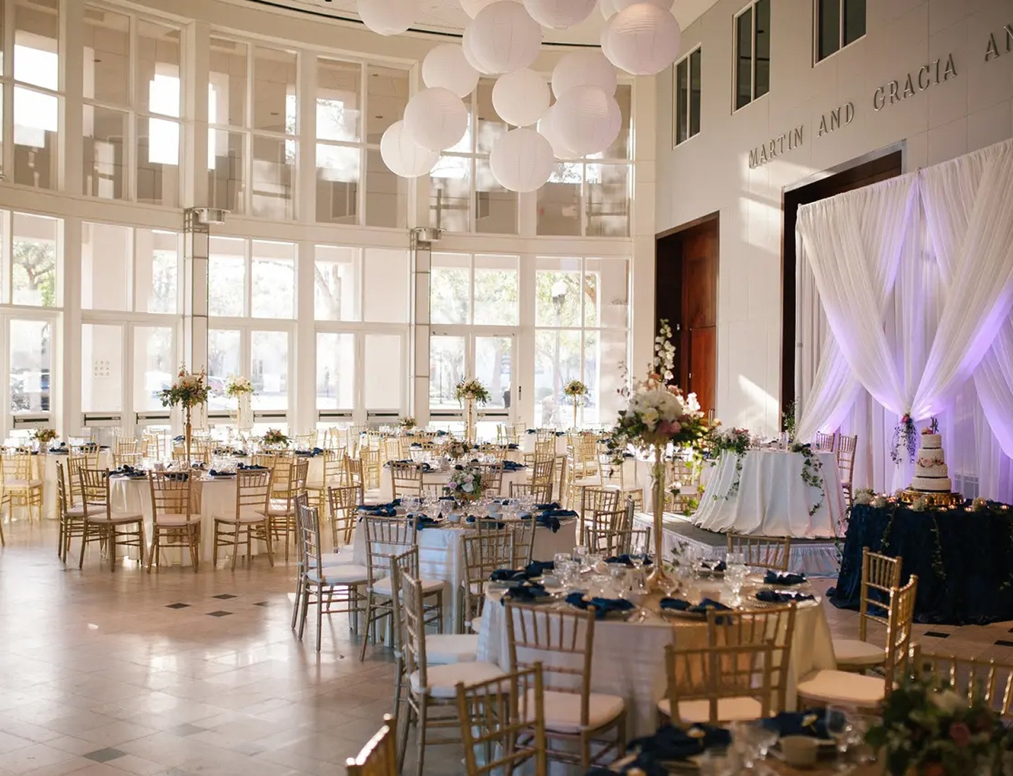 Airy event hall with gold chairs, large windows, tall florals, and white lanterns above the Orlando Museum of Art