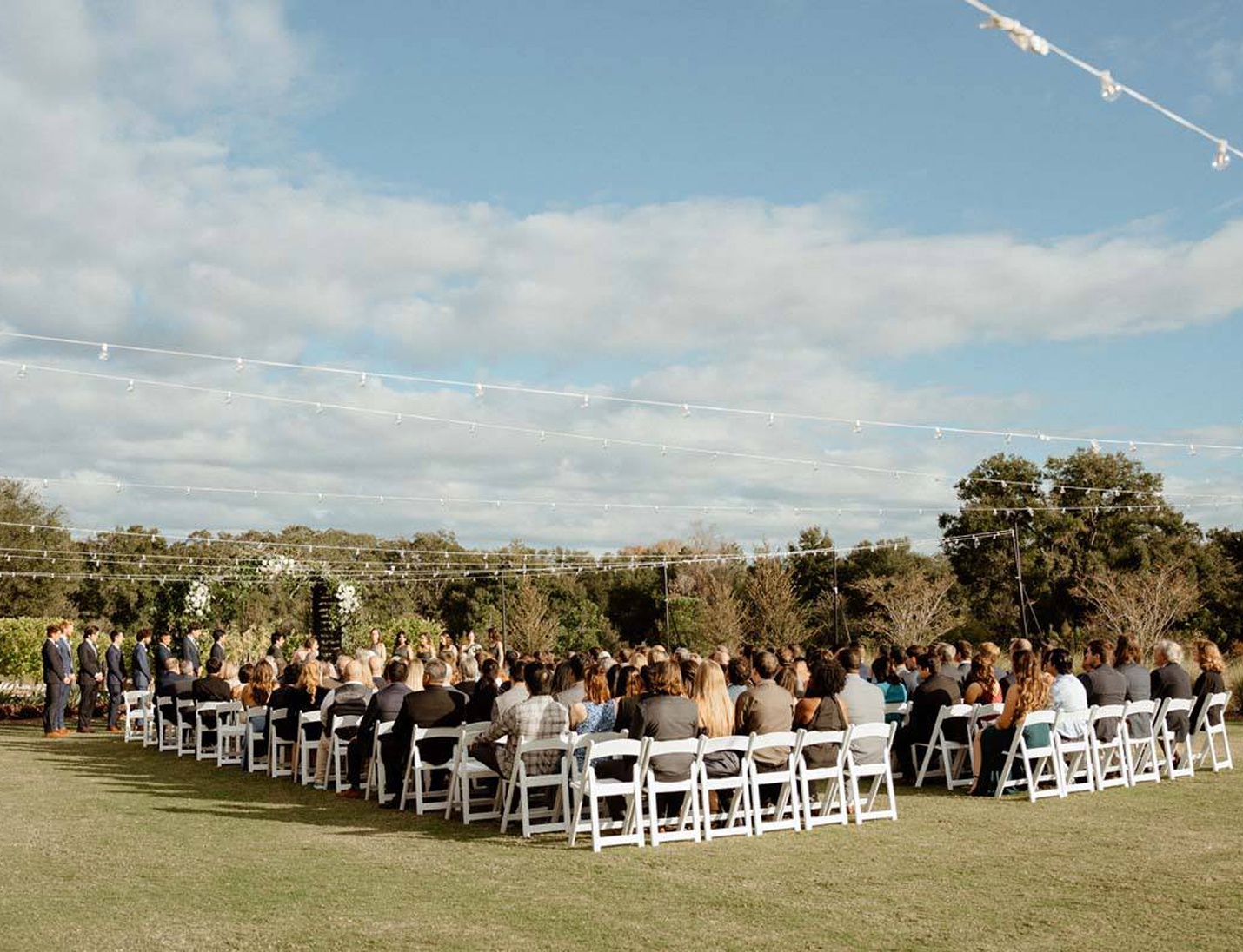 Outdoor wedding ceremony at Bonnet Springs Park with white chairs under string lights and a natural green backdrop