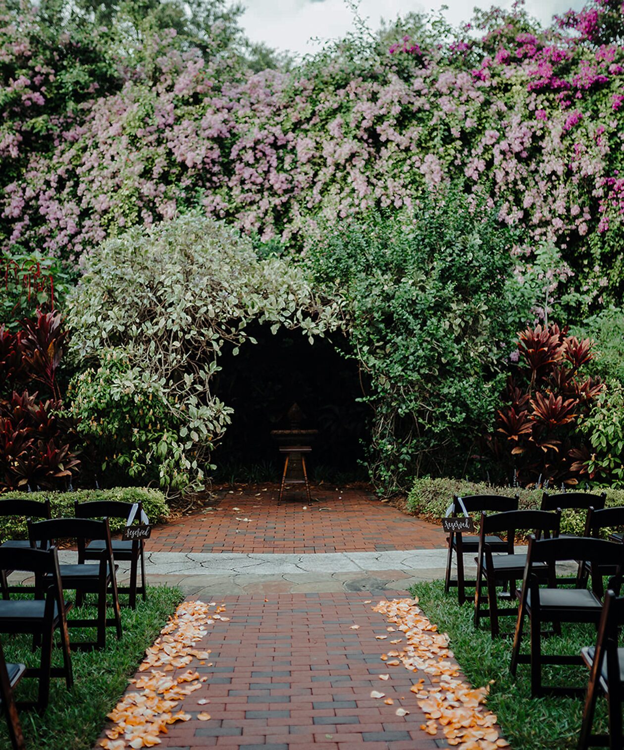 Garden ceremony setup at Sunken Gardens with a brick aisle, black chairs, lush greenery, and a blooming floral backdrop