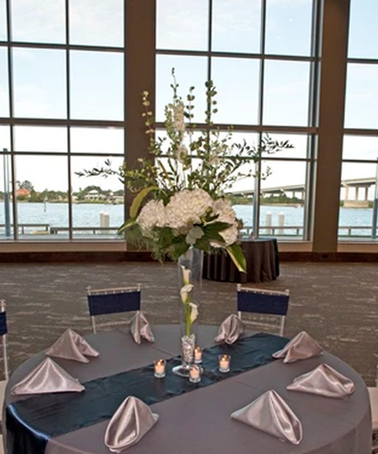 Elegant table setup with tall floral centerpiece and waterfront views through large windows at Brannon Civic Center