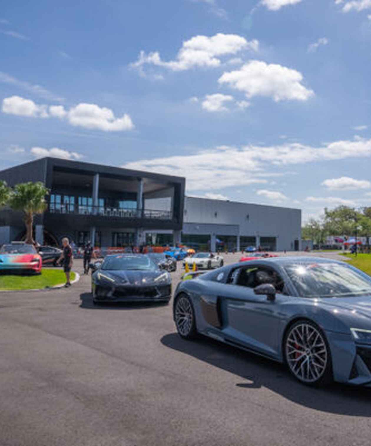 Exotic cars parked outside The Motor Enclave luxury automotive venue, with a modern building and a sunny outdoor setting