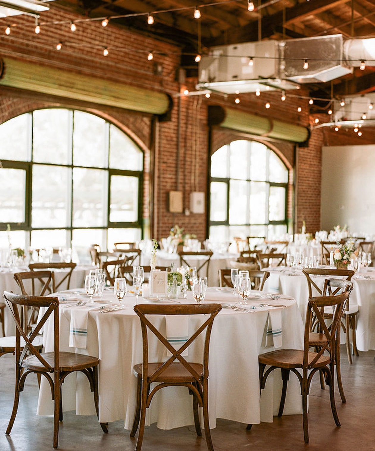 Warm brick interior with round tables, white linens, and wooden chairs under string lights at Morean Center for Clay