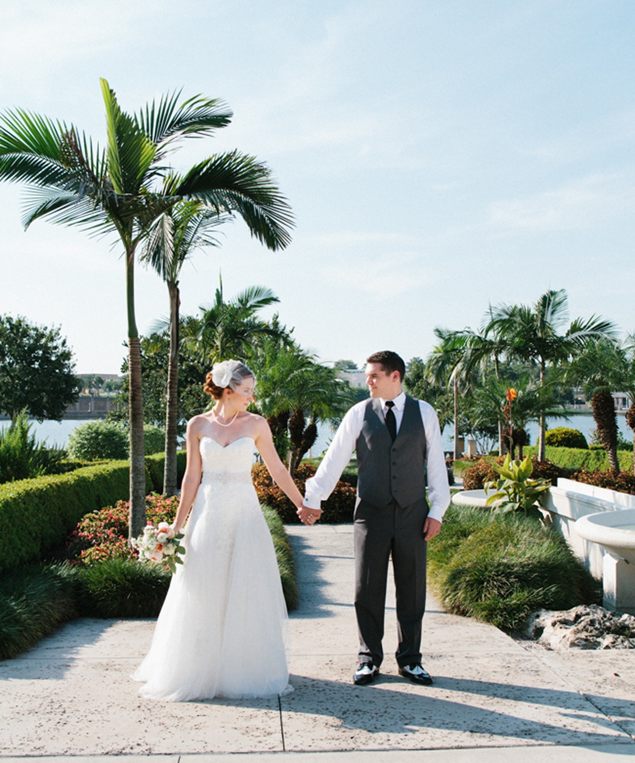 Bride and groom holding hands at Hollis Garden, surrounded by palm trees, and a peaceful waterfront backdrop
