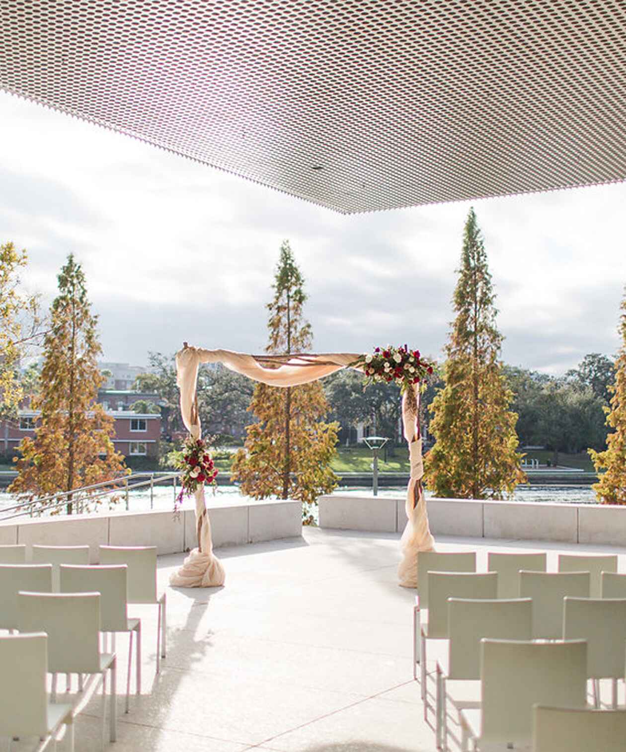 Outdoor wedding ceremony at Tampa Museum of Art with draped floral arch, modern seating and view of trees by the water