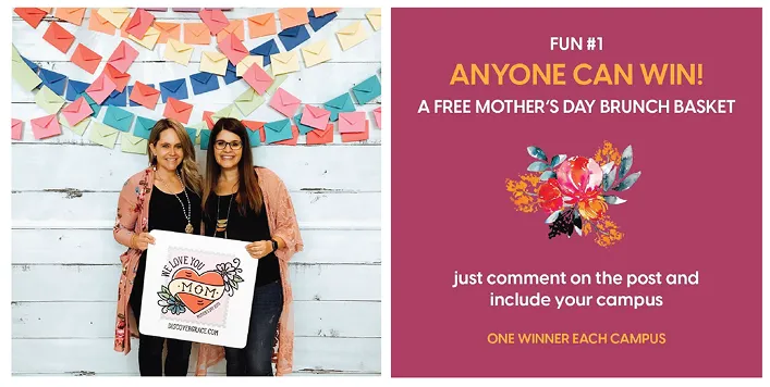 Two women smiling and holding a Mother’s Day sign in front of a colorful paper heart backdrop during an event promotion