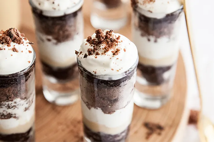 Layered dessert shooters with chocolate crumbs and whipped cream, arranged neatly on a wooden serving board