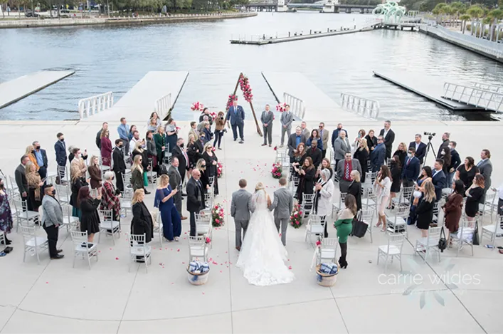Outdoor waterfront wedding ceremony at Armature Works, with guests gathered around the couple under a floral arch by the river