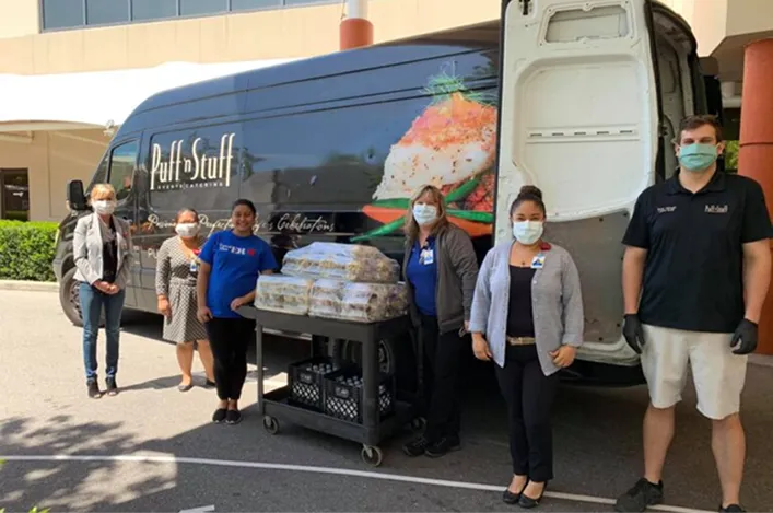 Team members standing beside a catering van, delivering packed meals with masks and gloves for safe community support