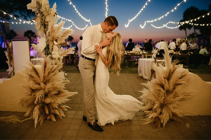 A couple sharing a romantic kiss surrounded by pampas grass decor under warm string lights at sunset