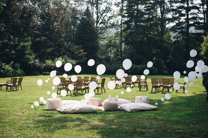 Outdoor lounge setup on a grassy lawn with pillows, poufs, and white floating balloons