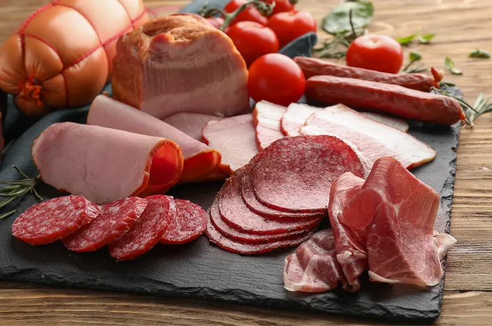 Variety of cured meats arranged on a slate board with sliced salami, ham, prosciutto, and tomatoes for serving