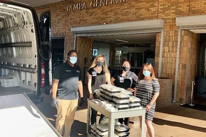 Team delivering packaged meals outside Tampa General Hospital, wearing masks and gloves, standing beside a service cart.
