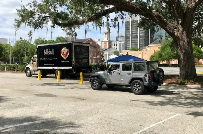 Puff’n Stuff Catering delivery truck and a parked Jeep in an outdoor lot with city buildings and trees in the background