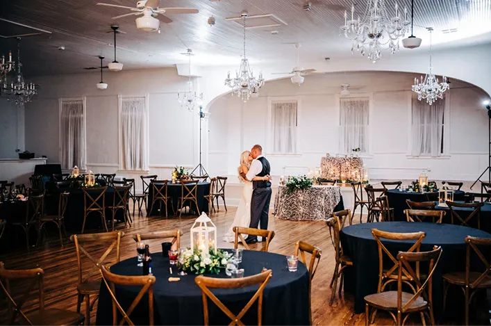 Bride and groom sharing a first dance in an elegant reception hall with chandeliers, round tables, and soft romantic lighting