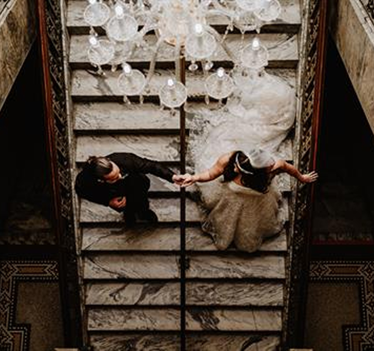 Bride and groom holding hands while descending a marble staircase beneath a chandelier at Centro Asturiano de Tampa
