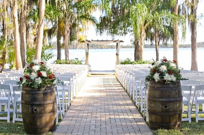 A lakeside wedding ceremony setup features rows of white chairs, wooden barrels with flowers, and a water backdrop