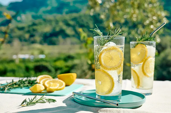 Tall iced lemon drinks garnished with rosemary, with sliced lemons and rosemary sprigs on an outdoor table by trees