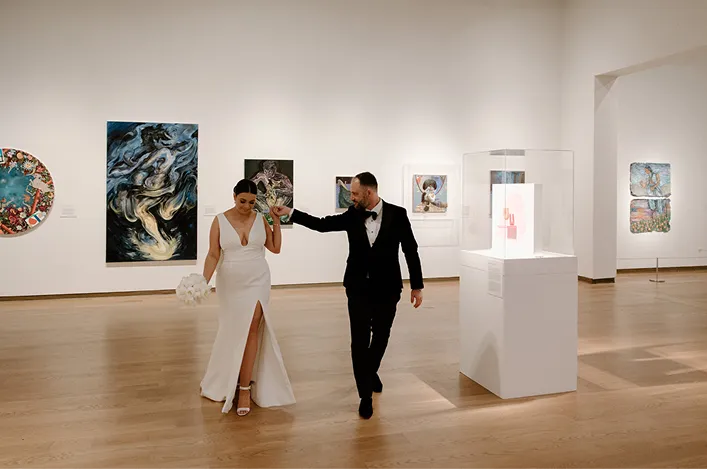 A bride and groom walk together in a modern art gallery with wood floors, abstract paintings, and elegant lighting