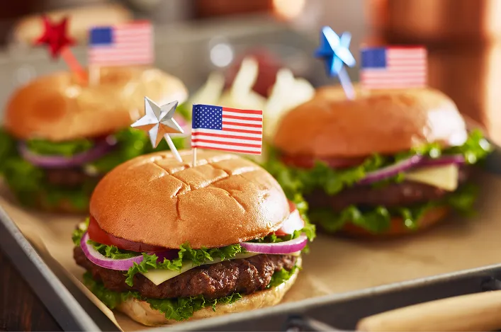 Classic cheeseburgers with lettuce, tomato, onion, and American flags on top are lined up on a tray for a celebration