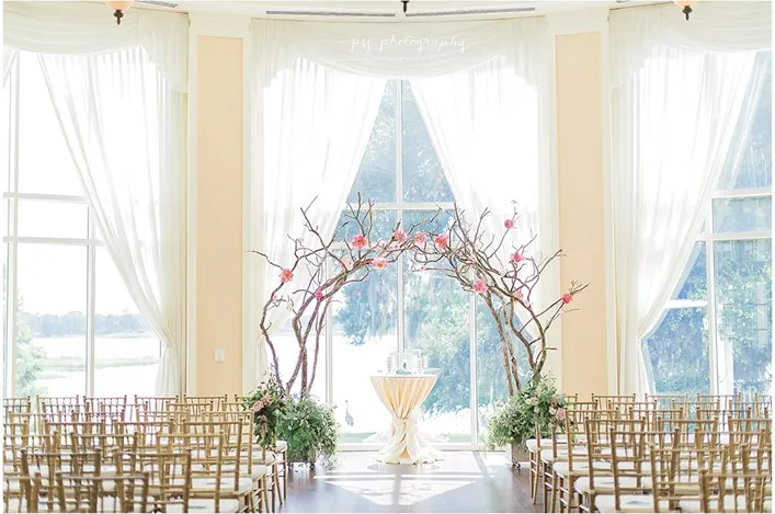 An indoor wedding ceremony space with gold chairs, a branch arch decorated with pink flowers, and tall sheer windows