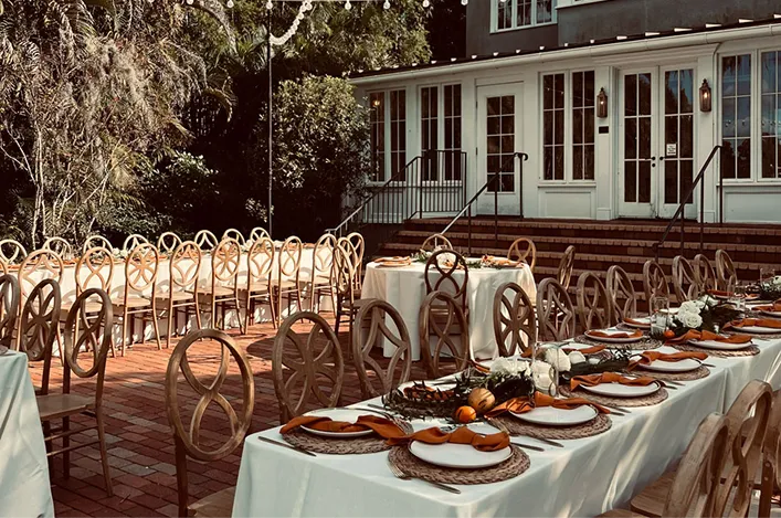 Elegant outdoor tables are set with white linens, wooden chairs, orange napkins, and minimalist centerpieces for a garden event