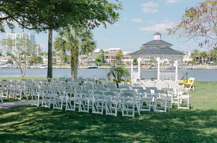 Rows of white chairs are arranged on green grass facing a gazebo beside a lake, ready for an outdoor ceremony