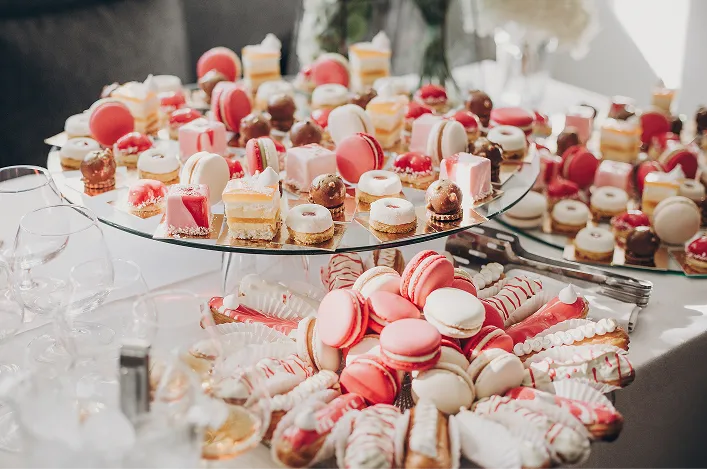 A dessert table is filled with colorful macarons, petit fours, and assorted mini pastries arranged on glass trays for an event