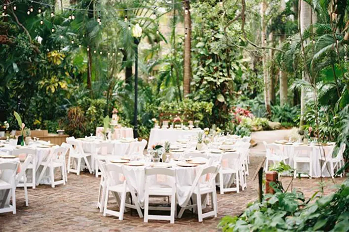 Outdoor garden venue with round tables, white chairs, and string lights surrounded by lush greenery and tropical plants