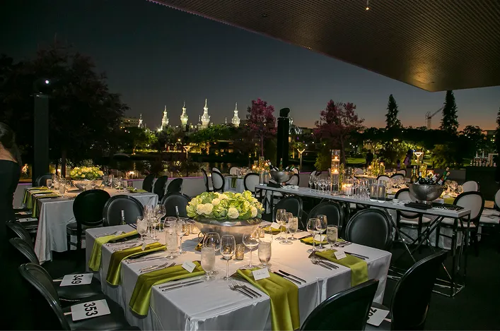 Elegant outdoor evening reception setup with white linens, green napkins, candlelit centerpieces, and city skyline in the background