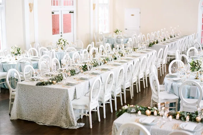 Elegant indoor reception setup with long tables, white chairs, soft linens, greenery garlands, and simple floral centerpieces