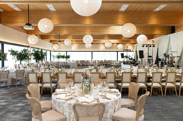 Bright reception hall with round tables, neutral linens, elegant place settings, globe pendant lights, and a live band setup